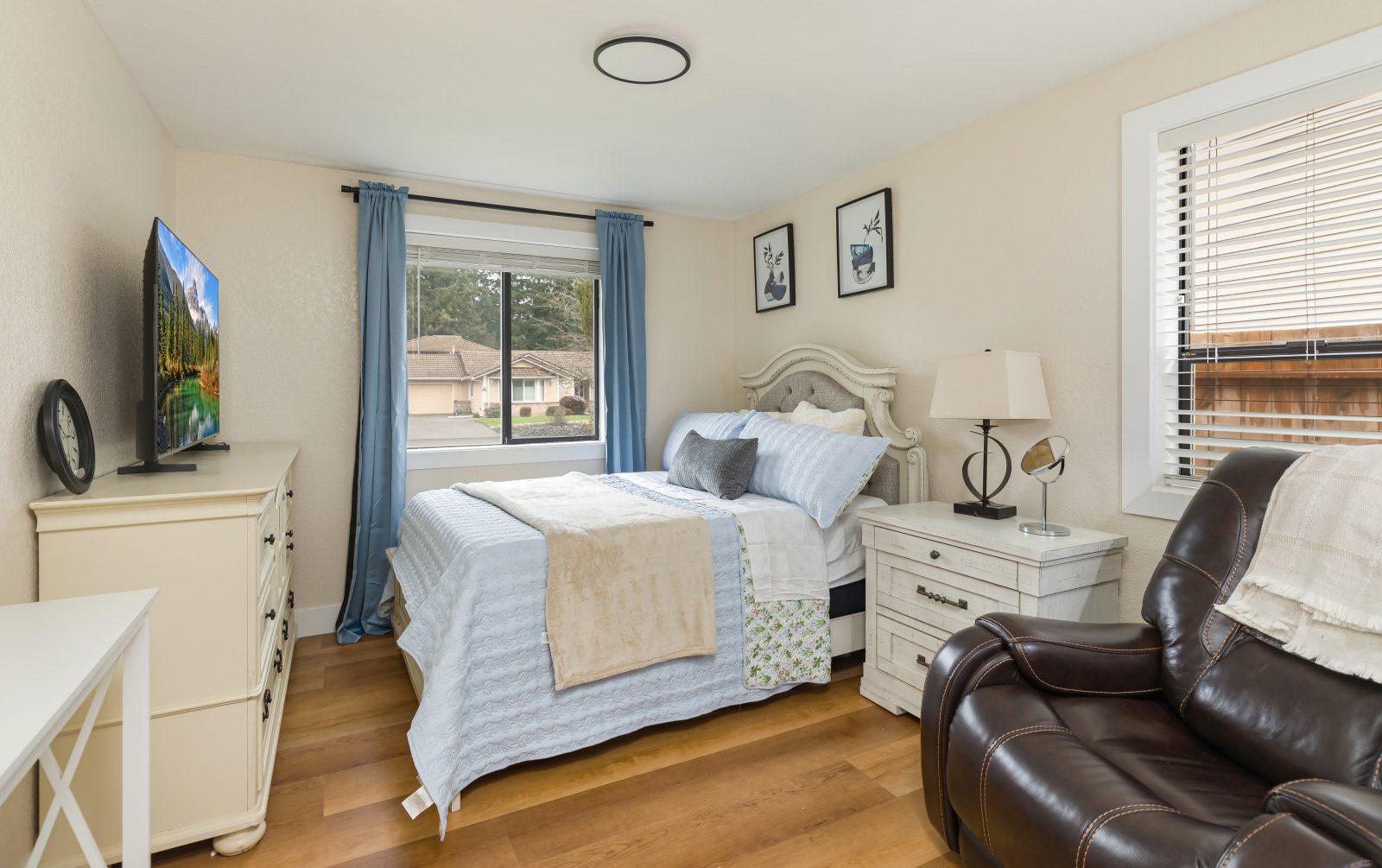A private bedroom with a bed, dresser, recliner and two bright windows.